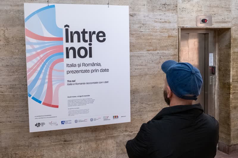 A visitor in a blue cap reads the Între noi title panel beside a lift.