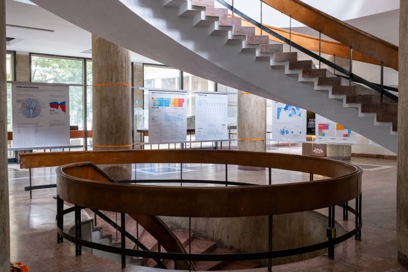 The exhibition seen beneath a curving marble staircase, panels strung between the columns.